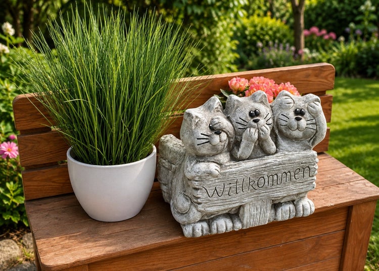 Steinfigur drei Katzen mit Willkommen Schild als Gartendekoration auf einer Holzbank stehend neben einem Blumentopf mit Gras