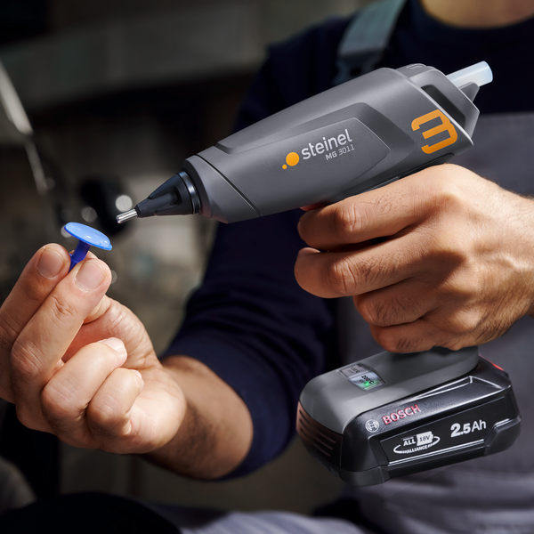 Eine Person hält eine kabellose Steinel Heißklebepistole MG 3011 mit Bosch Akku beim Arbeiten mit einem Klebepin in der Hand.