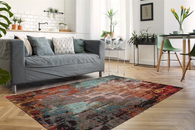 Heller, moderner Wohnraum mit abstrakt gemustertem Teppich auf Parkettboden, grauem Sofa mit Kissen und einem Essbereich mit Pflanzen.