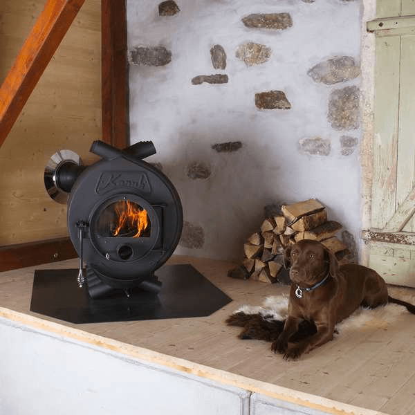 Rustikaler Innenraum mit rundem Werkstattofen aus Metall, Kanuk Logo, brennendem Feuer, Brennholzstapel und Hund auf Holzboden vor Steinwand.