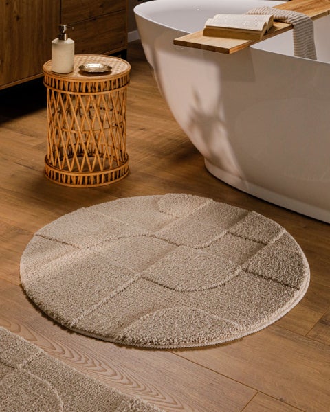 Runde Badematte mit Reliefmuster auf Holzboden neben freistehender Badewanne und Rattan-Beistelltisch in einem hellen, modernen Badezimmer.
