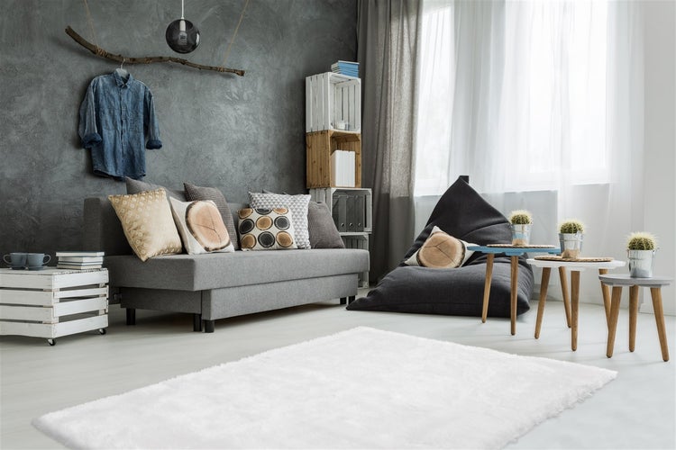 Modernes Wohnzimmer mit grauem Sofa, Sitzsack, strukturierter grauer Wand, weißem Teppich und Beistelltischen aus Holz.