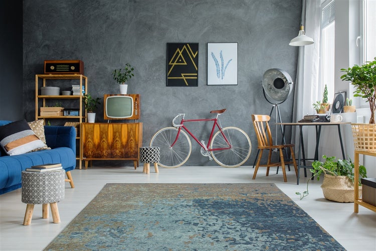 Wohnzimmer mit grau strukturierter Wand, blauem Sofa, Vintage-Möbeln, Rennrad und einem abstrakten Teppich in Blau- und Goldtönen.