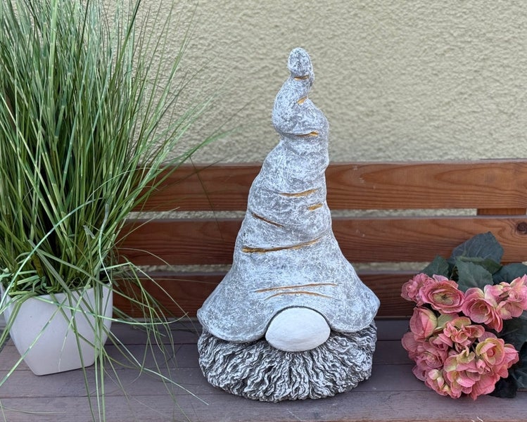 Deko-Gartenzwerg aus Stein mit großer Zipfelmütze und Bart, platziert auf einer Holzbank neben einer Topfpflanze und Blumen.