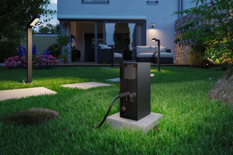 Moderne Gartensteckdosensäule auf einem Betonfundament in einem beleuchteten Garten vor einer Terrasse am Abend.