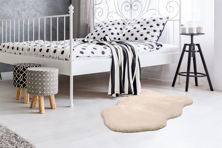 Helles Schlafzimmer mit weißem Metallbett, Bettwäsche mit Punkten, zwei gemusterten Hockern und einem beigen Kunstfellteppich.