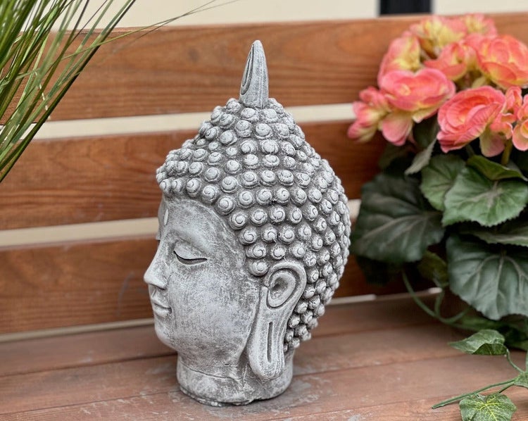 Dekorative Buddha-Kopf Gartenfigur aus Steinguss auf einer Holzoberfläche neben einer Pflanze