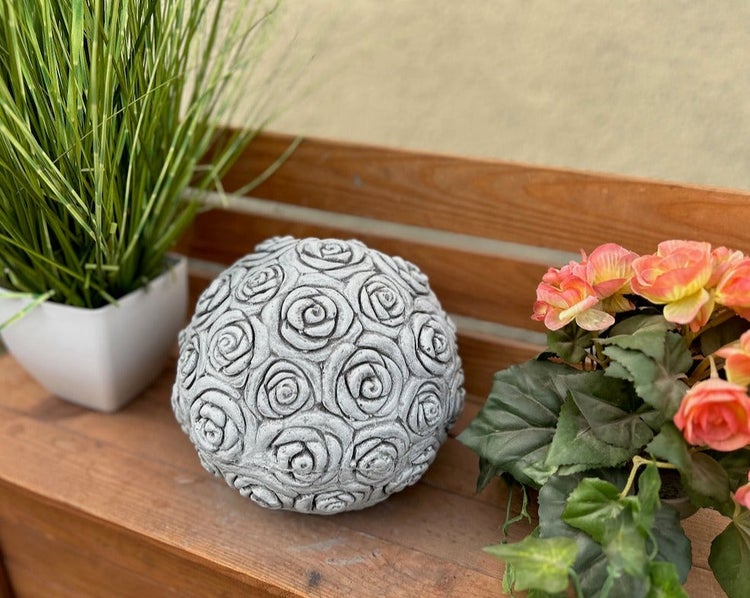 Dekorative Gartenkugel aus Beton mit Rosen-Muster auf einer Holzbank, neben einer Grünpflanze und Blumen.