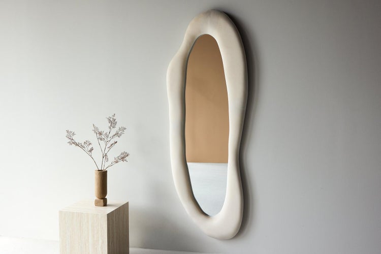 Asymmetrischer Wandspiegel mit breitem, wellenförmigem Rahmen im organischen Design, neben einem Holzsockel mit Trockenblumen.