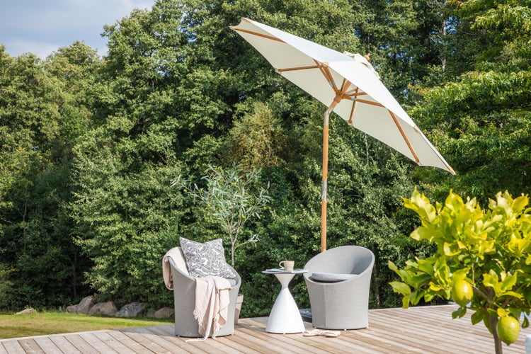 Großer weißer Sonnenschirm mit Holzgestell über grauen Gartensesseln und weißem Beistelltisch auf einer Holzterrasse vor einem Wald.