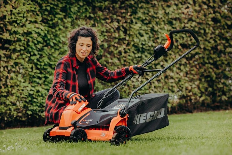Frau bedient einen Rasenmäher der Marke Echo auf einer Rasenfläche vor einer Hecke.