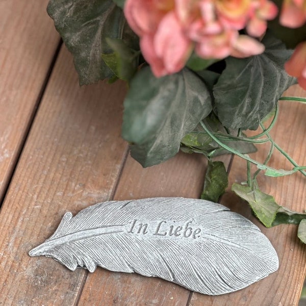 Grabschmuck Feder aus Kunststein mit der Aufschrift In Liebe, platziert neben einer Pflanze.