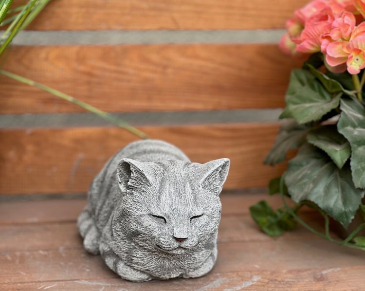Gartenfigur schlafende Katze in Steinoptik platziert auf einer Holzbank vor Holzzaun mit Blumen.