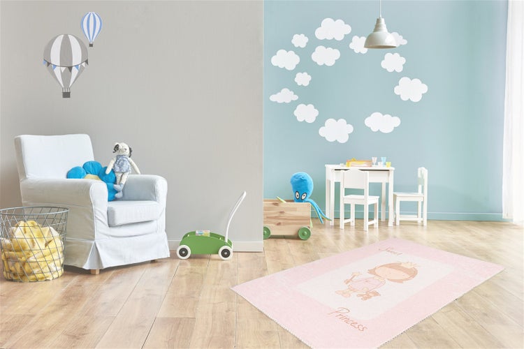 Helles Kinderzimmer mit Kindersessel, Spielzeug und einem rosa Kinderteppich mit Prinzessinnen-Motiv vor einer blauen Wand mit Wolken-Wandstickern.
