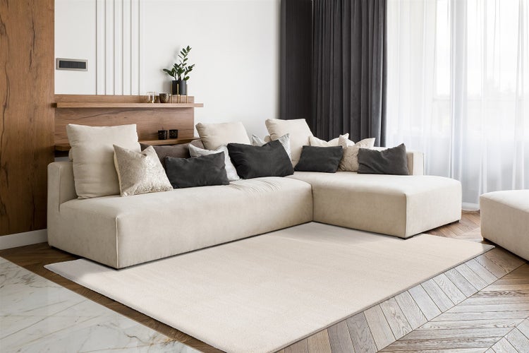 Modernes Wohnzimmer mit cremefarbenem Ecksofa, hellem rechteckigem Teppich auf Parkettboden und dunklen Vorhängen. Helle Atmosphäre.