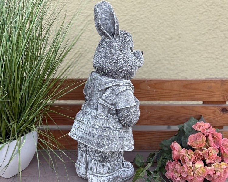 Gartenfigur Hase in Steinoptik, bekleidet mit Kleid und Stiefeln, platziert auf einer Holzbank neben Pflanzen.