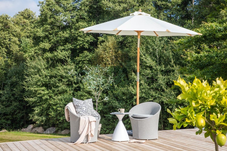 Sonnenschirm aus Holz mit weißem Stoff, zwei graue Loungesessel mit Kissen und kleiner weißer Beistelltisch auf einer Holzterrasse vor Wald.