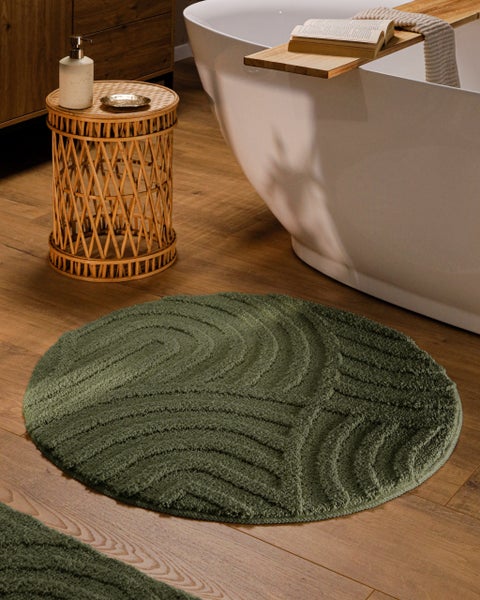 Runder grüner Badteppich mit strukturiertem Wellenmuster auf Holzfußboden neben einer weißen Badewanne und einem Beistelltisch aus Rattan.