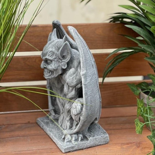 Wasserspeier Gartenfigur aus Stein in Form eines Dämons stehend auf einem Sockel zwischen Pflanzen