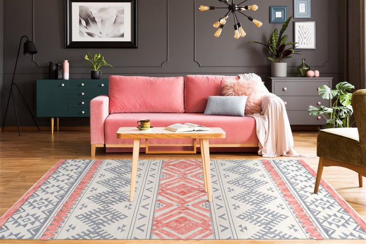 Modernes Wohnzimmer mit rosa Samtsofa, hölzernem Couchtisch und geometrisch gemustertem Teppich vor einer dunkelgrauen Wand mit Zierleisten.