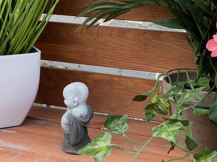 Kleine Buddha Gartenfigur aus Stein auf einer Holzbank zwischen Topfpflanzen als Dekoration für den Außenbereich