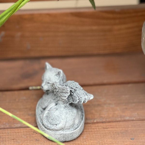 Steinfigur einer schlafenden Katze mit Engelsflügeln als Grabschmuck oder Gartendeko auf einem Holzuntergrund.