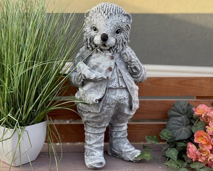Dekorative Gartenfigur Igel in Steinoptik mit Kleidung, stehend auf einer Holzbank neben einer Topfpflanze.