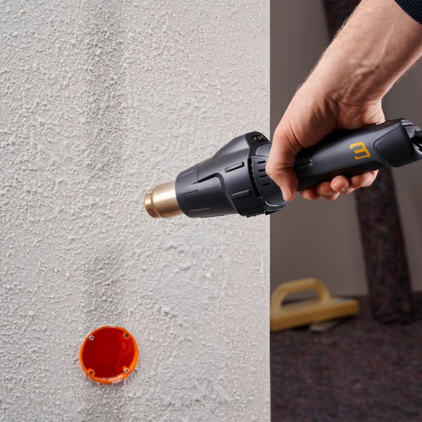 Eine Hand hält eine Heißluftpistole vor eine verputzte Wand mit einer Unterputzdose für Elektroinstallationen.