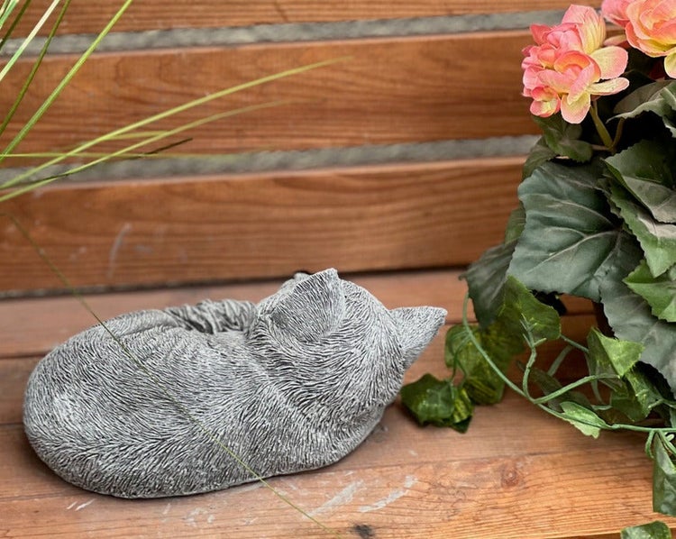 Gartenfigur einer schlafenden Katze in Steinoptik auf einer Holzoberfläche neben Pflanzen.
