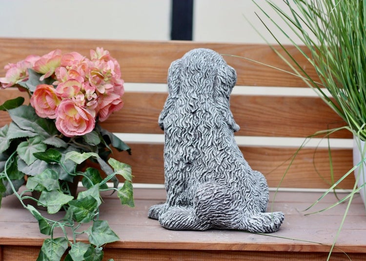 Gartenfigur sitzender Hund in Steinoptik von hinten auf einer Holzbank neben Blumen.