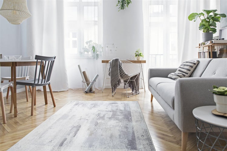Helles Wohnzimmer mit grauem Sofa, Holztisch und abstrakt gemustertem Teppich auf Fischgrätenparkett.