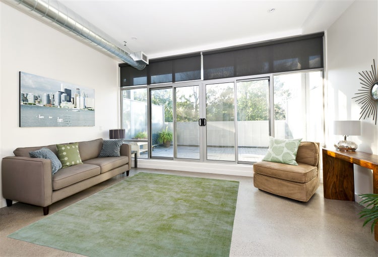 Hell gestaltetes Wohnzimmer mit Sofa, Teppich, Sessel und großen Glasschiebetüren zum Balkon. Über der Fensterfront sind Rollos montiert.