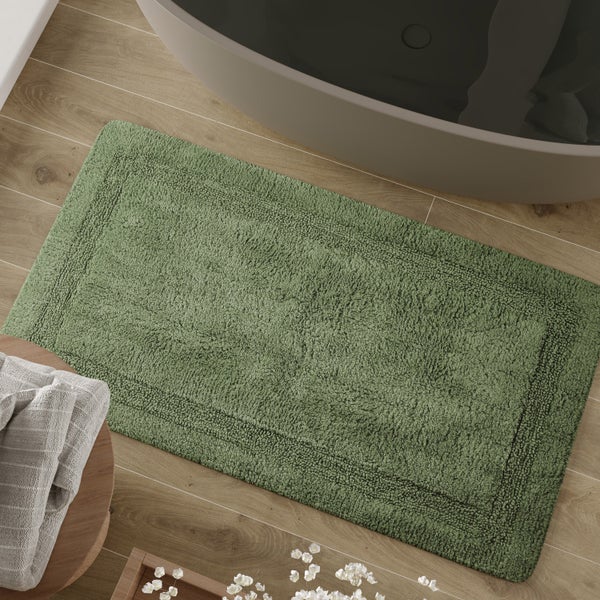 Rechteckige Badematte mit gewebter Bordüre auf einem Boden in Holzoptik neben einer modernen Badewanne.