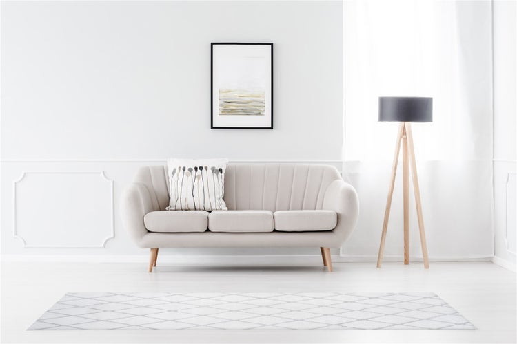 Helles, modern eingerichtetes Wohnzimmer mit beigem Sofa, Dreibein-Stehleuchte aus Holz und einem Teppich mit Rautenmuster.