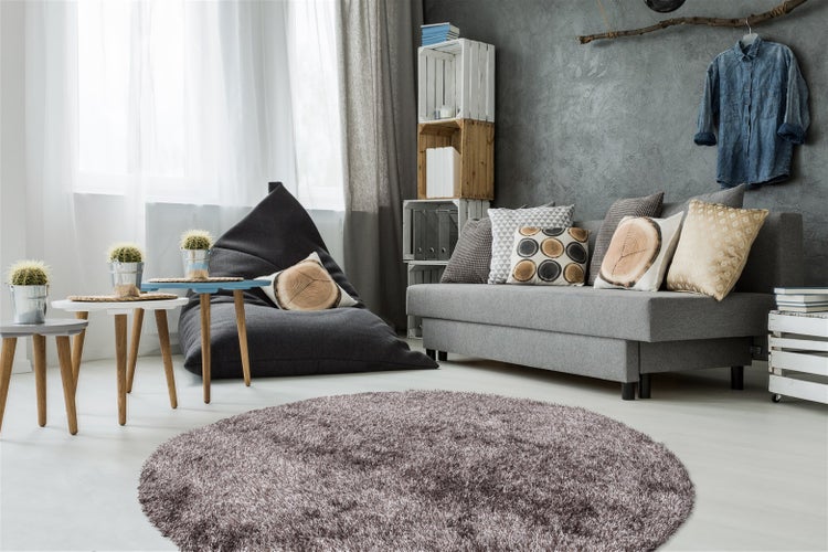 Runder Hochflor-Teppich in einem modern eingerichteten hellen Wohnzimmer mit grauem Sofa, großem Sitzsack und Beistelltischen aus Holz.
