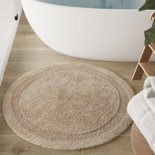 Runder Badeteppich in Beige vor einer weißen Badewanne im Badezimmer auf einem Boden in Holzoptik.