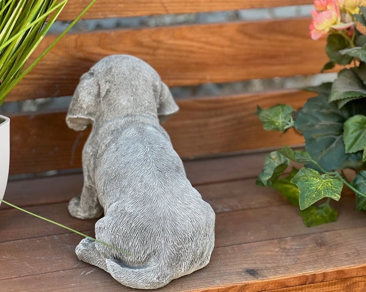 Gartenfigur Hund in Steinoptik von hinten sitzend auf einer Holzbank mit Pflanzen.