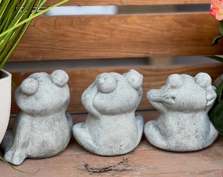 Drei sitzende Frosch Gartenfiguren aus Steinguss in unterschiedlichen Posen vor einem Holzlattenzaun.