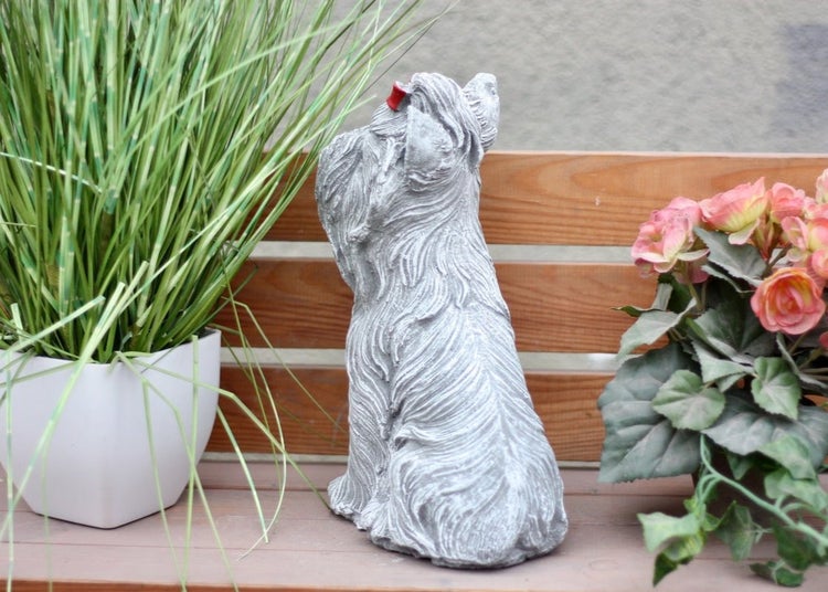 Gartenfigur Yorkshire Terrier aus Steinguss, sitzend von hinten mit roter Schleife im Haar, platziert auf einer Holzbank zwischen Pflanzen.