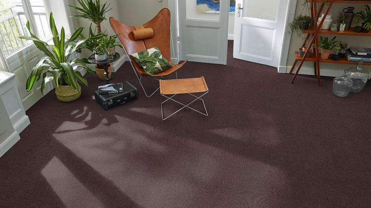Dunkelbraune Teppichfliesen mit Rippenstruktur in einem hellen Wohnzimmer mit braunem Ledersessel und grünen Zimmerpflanzen.