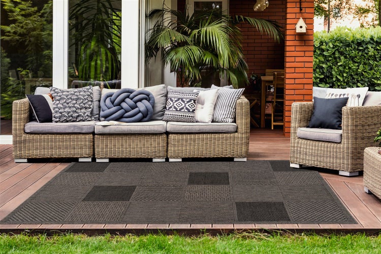 Grauer Outdoor-Teppich mit Patchwork-Muster auf einer Holzterrasse mit Polyrattan-Gartenmöbeln und gemusterten Kissen.