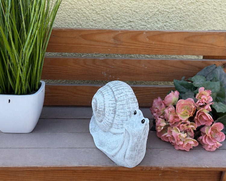Dekorative Schneckenfigur aus Stein für den Garten, auf einer Holzbank mit Pflanze und Blumen platziert.