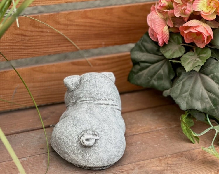 Gartenfigur aus Stein in Form eines Schweins von hinten, platziert auf Holzbrettern neben einer Pflanze.