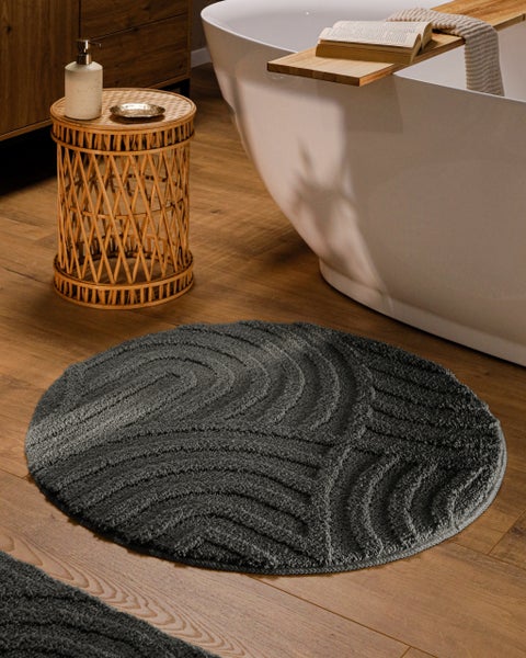 Runde graue Badematte mit Wellenstruktur in einem hellen Badezimmer neben einer freistehenden Badewanne auf Holzboden.