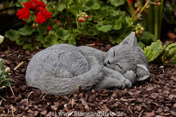 Eine liegende Gartenfigur in Form einer schlafenden Katze aus Stein im Gartenbeet.