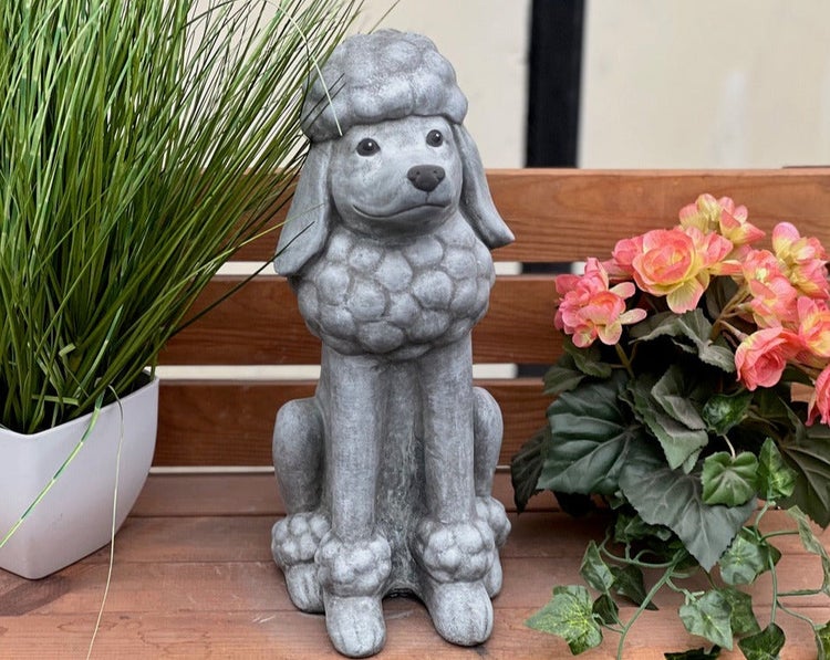 Steinfigur eines sitzenden Pudels als Gartendekoration auf einer Holzbank, flankiert von Pflanzen.