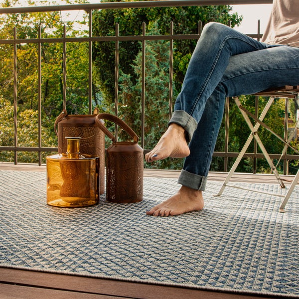 Gemusterter Outdoor-Teppich auf einem hellen Balkon mit dekorativen Laternen, einer Glasvase und einer sitzenden Person.