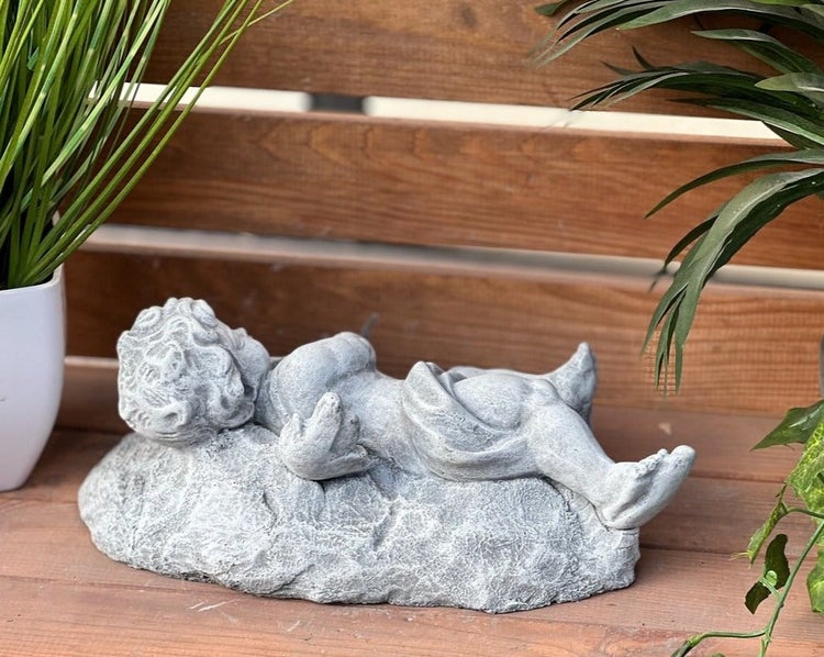 Liegende Putten Figur aus Stein für die Gartendekoration vor einer hölzernen Wand mit Pflanzen.