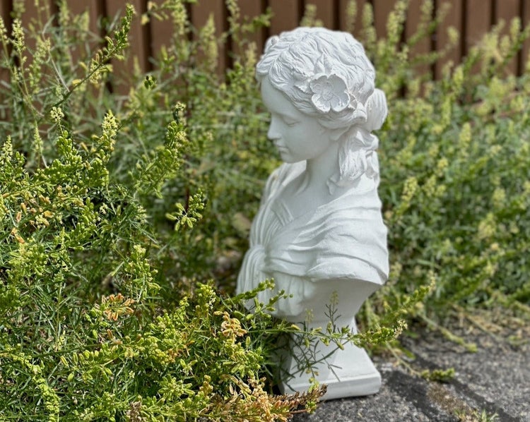 Weiße Gartenbüste einer jungen Frau mit Blumen im Haar, platziert inmitten von grünen Gartenpflanzen.
