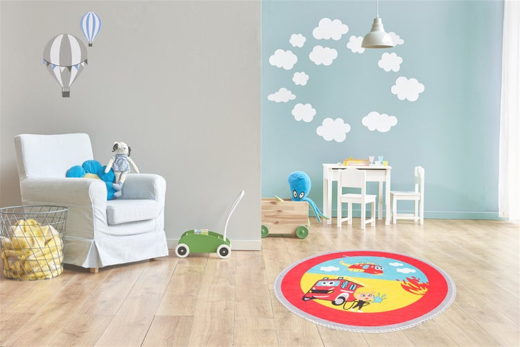 Runder Kinderteppich mit Feuerwehrmotiv in einem hellen Kinderzimmer mit Holzboden, weißem Sessel und Kindermöbeln.